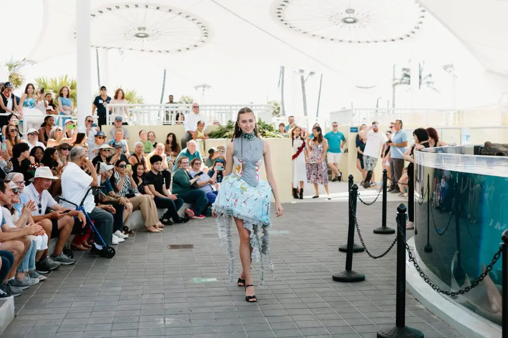 young woman in blue and white dress walks by a crowd seated in an amphitheater on the left and a large outdoor aquarium on the right.